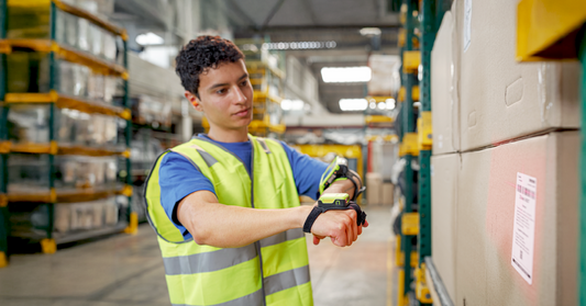 Person in a warehouse wearing a safety vest and using a handheld device.